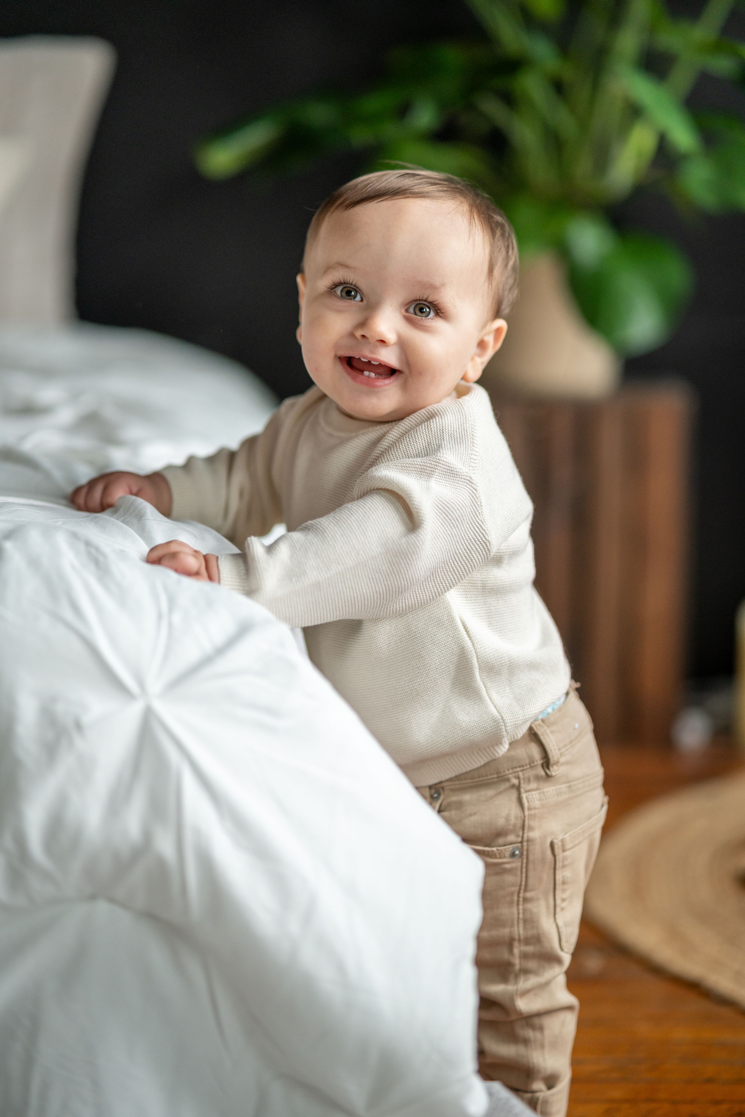 Smiling baby in white sweater standing by bed with houseplant in background.