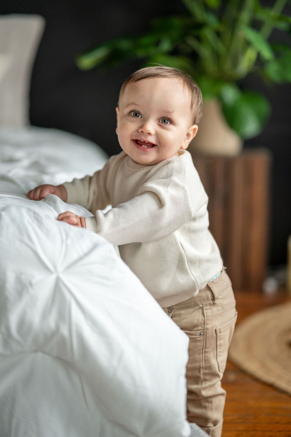 Smiling baby in white sweater standing by bed with houseplant in background.