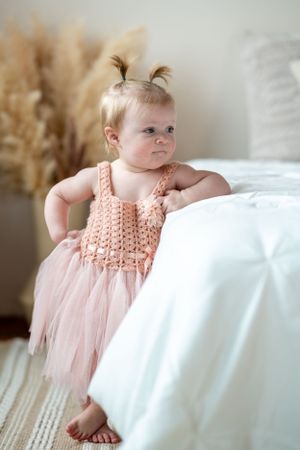 Baby standing by white bedding wearing peachy pink tulle dress with crochet top.