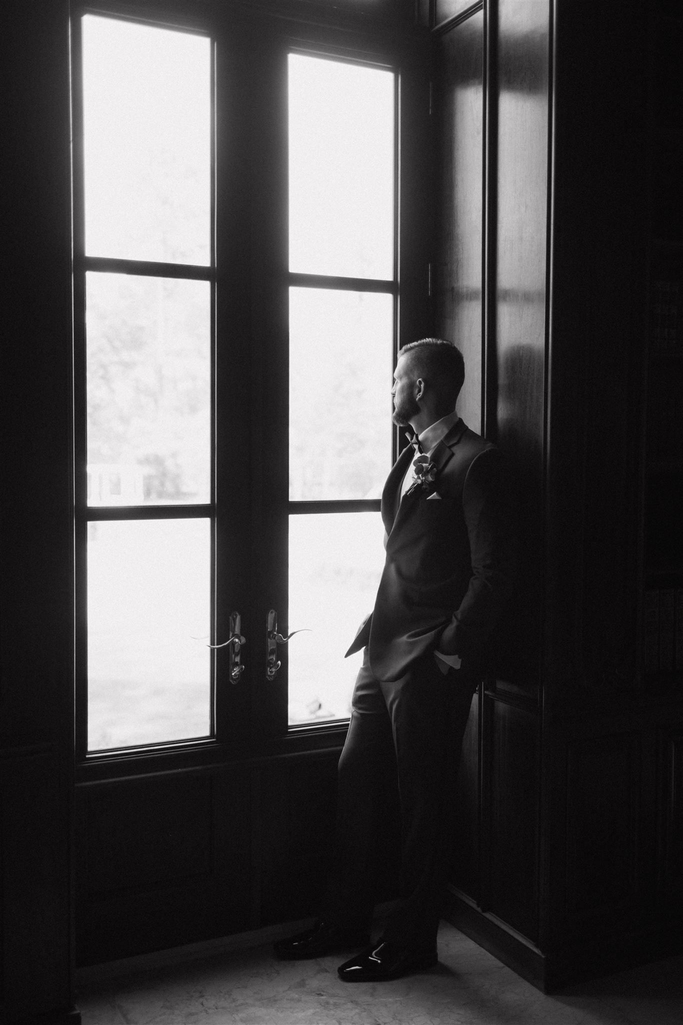 A person in a dark suit gazes contemplatively out tall windows in a moody black and white photograph.
