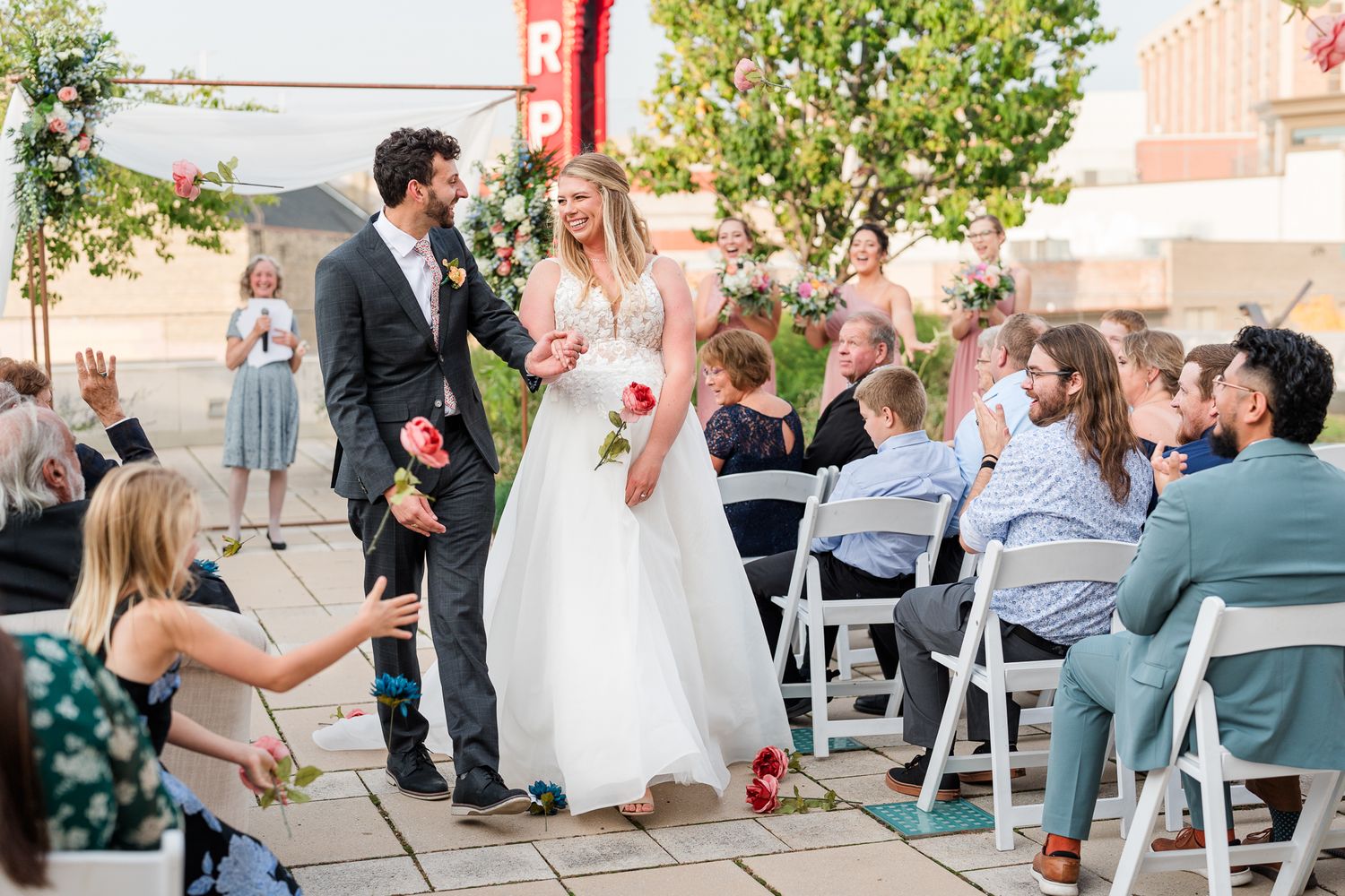 Wedding guests enjoy an outdoor celebration under trees with white chairs and decorative elements.