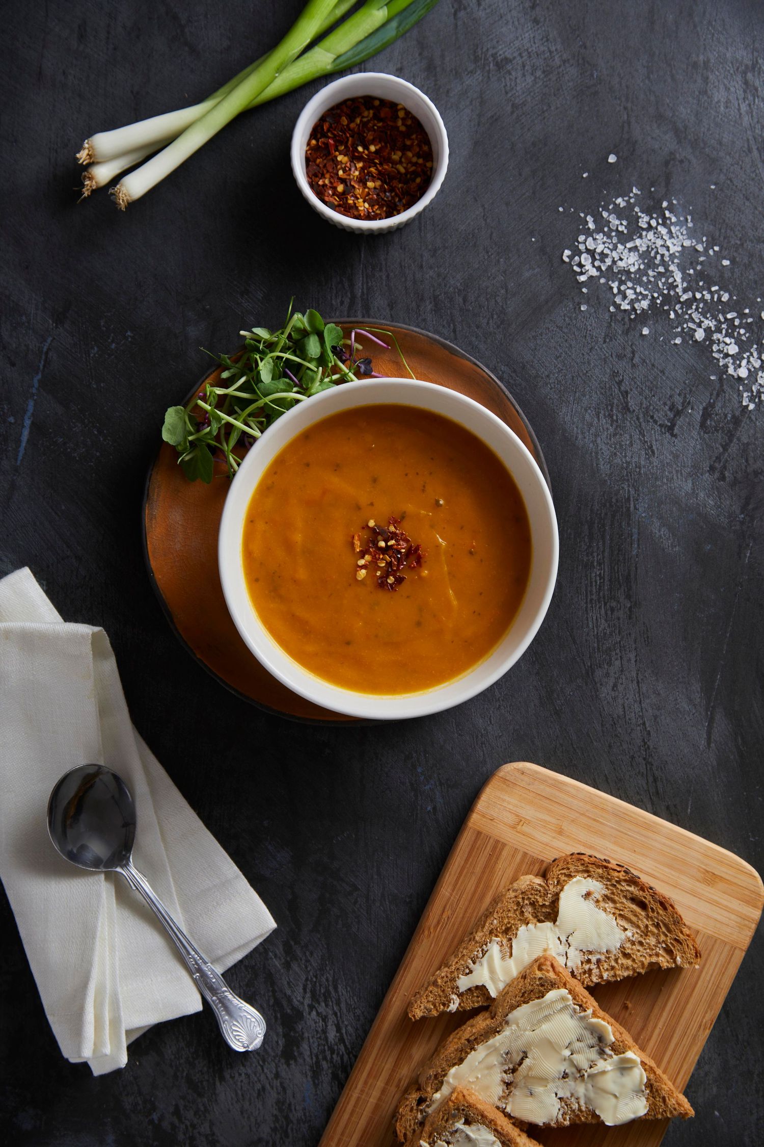 flatlay image of chilli soup in white bowl with watercress on the side a wooden board with granary buttered bread spoon