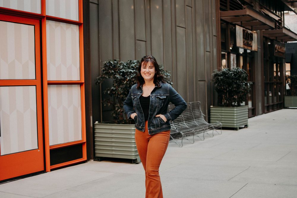A person in a denim jacket and orange pants poses confidently on a city sidewalk near an orange-framed door.