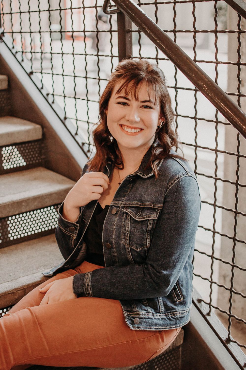A person wearing a denim jacket and orange pants poses on metal stairs with an industrial backdrop.