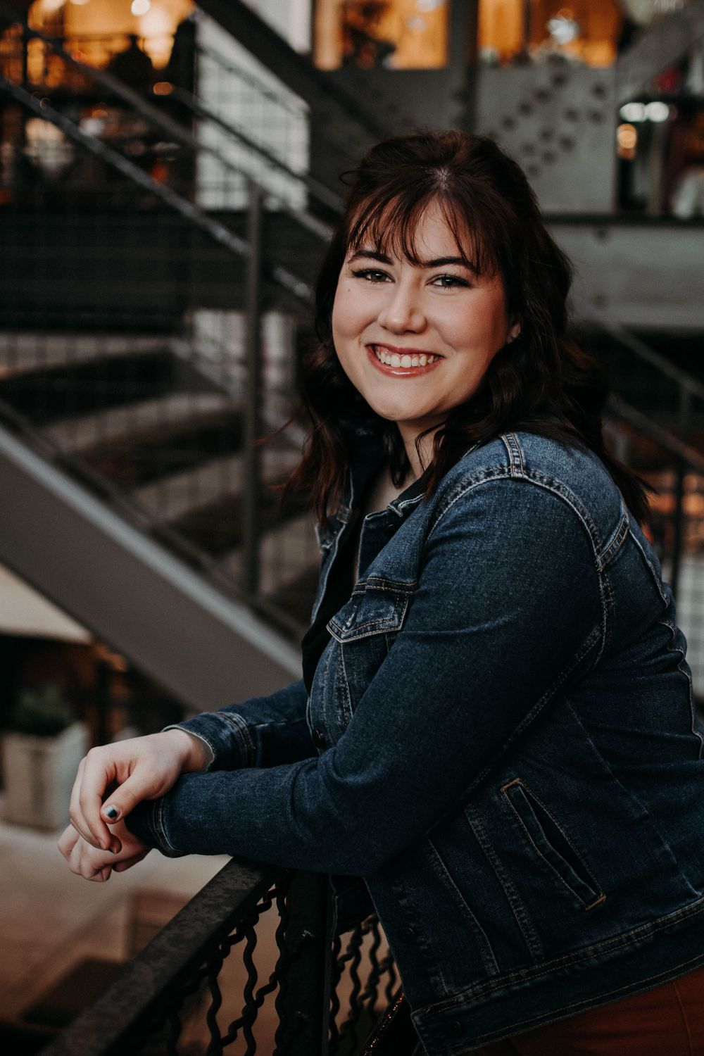 A person in denim poses by metal railings with warm interior lighting visible in the background.