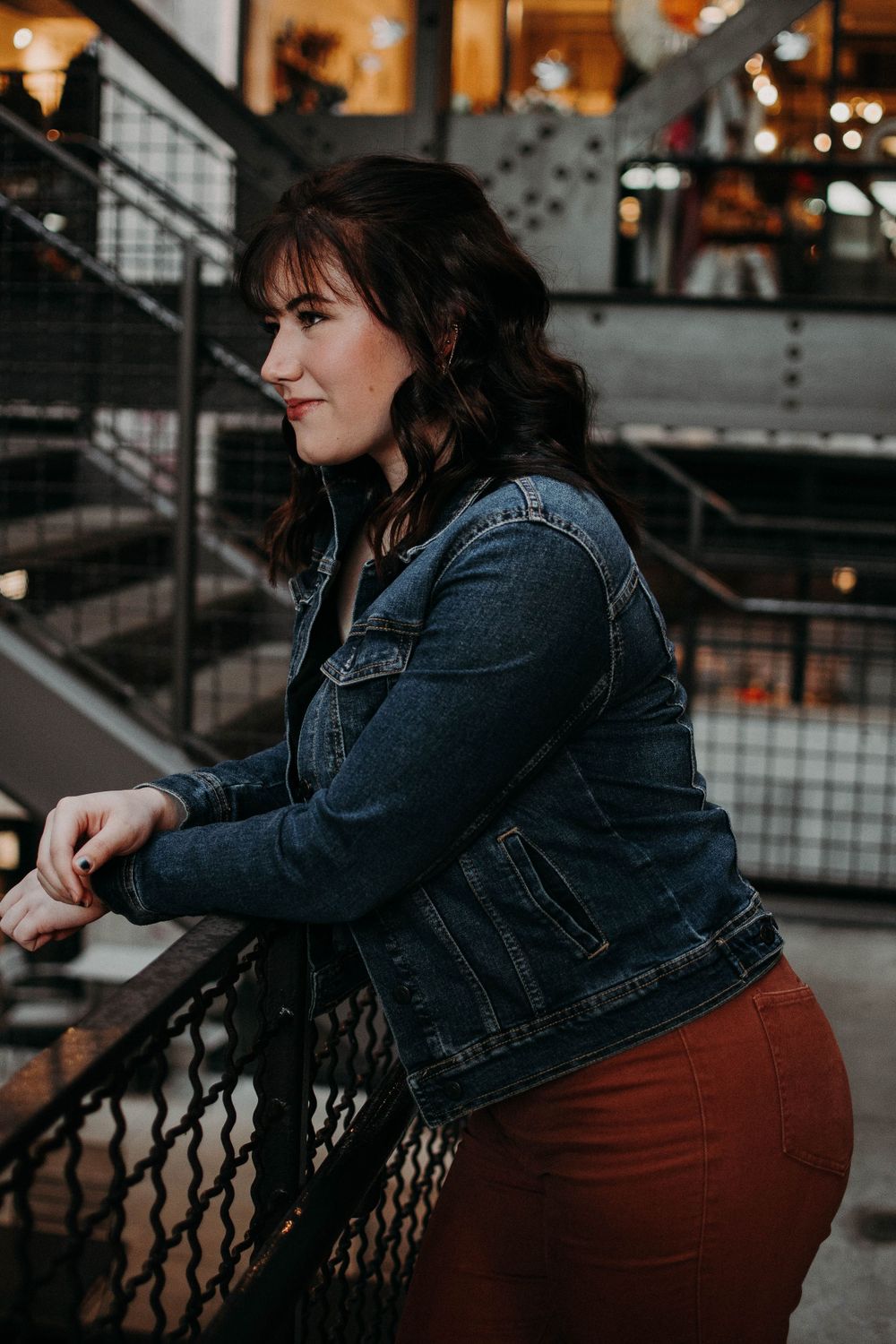 A person in a denim jacket leans on metal railings with warm lighting and industrial elements in view.