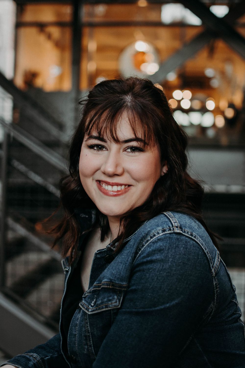 A person in denim smiles warmly against an industrial backdrop with warm lighting.
