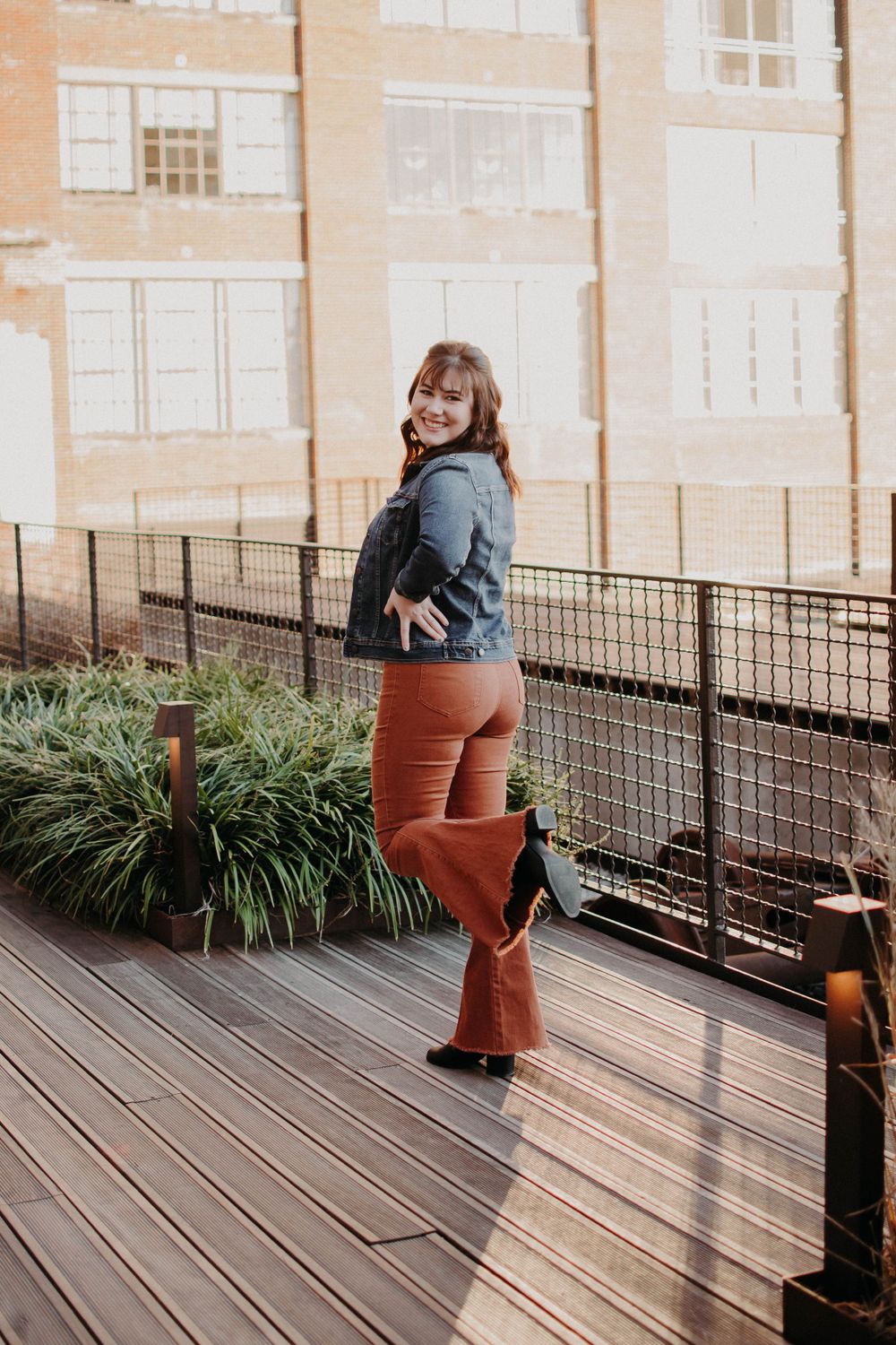 A person in denim and orange pants poses on a wooden deck with architectural elements in the background.
