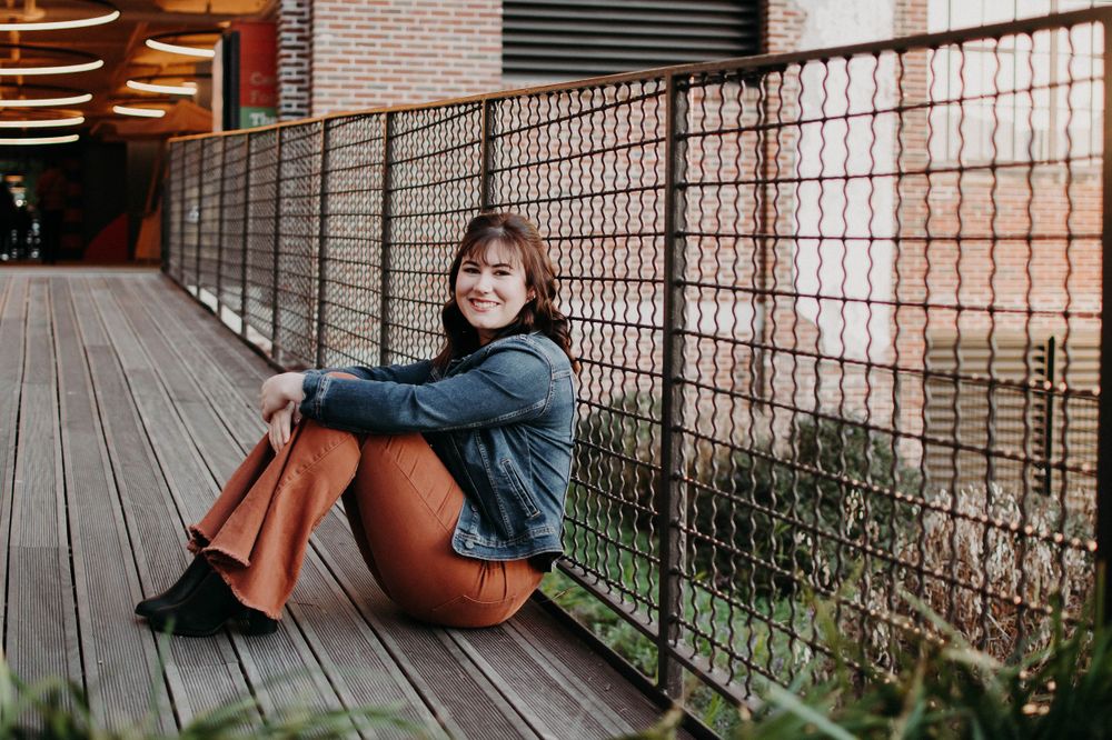 A casual portrait of a person sitting on wooden decking against metal railings and brick walls.