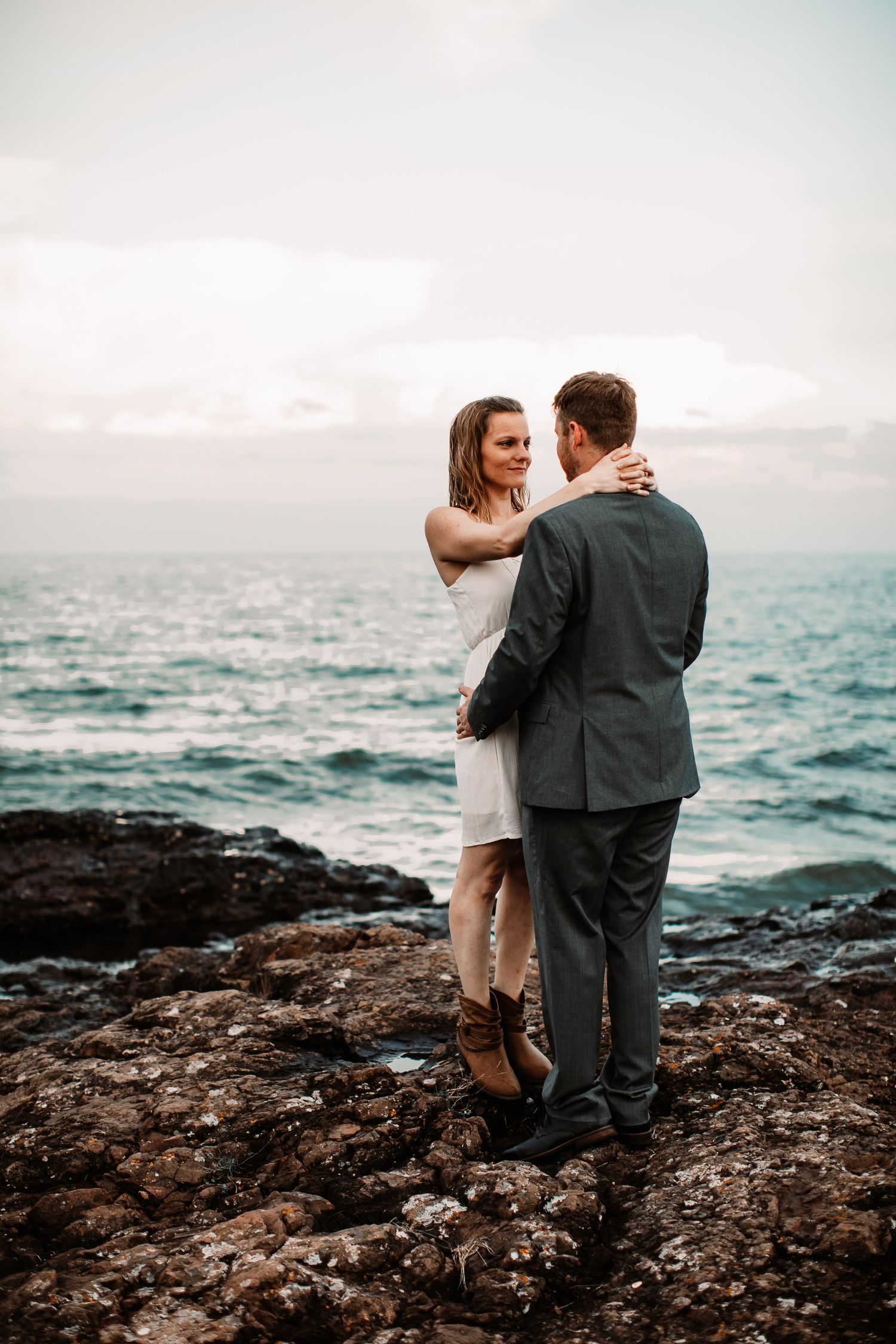 Couple embracing on rocky cliff overlooking ocean at sunset.