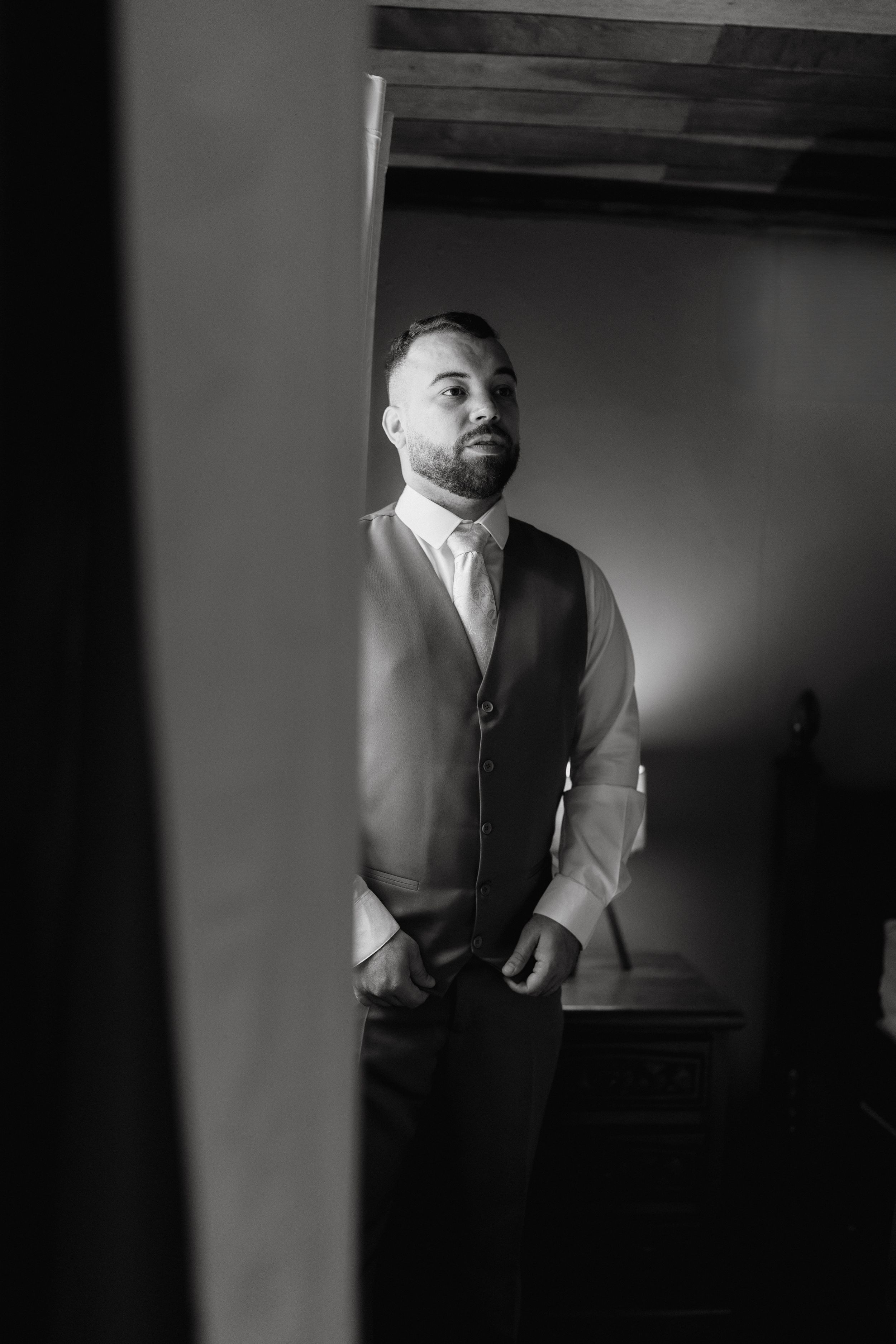 Black and white portrait of a groom in formal attire standing in a room.