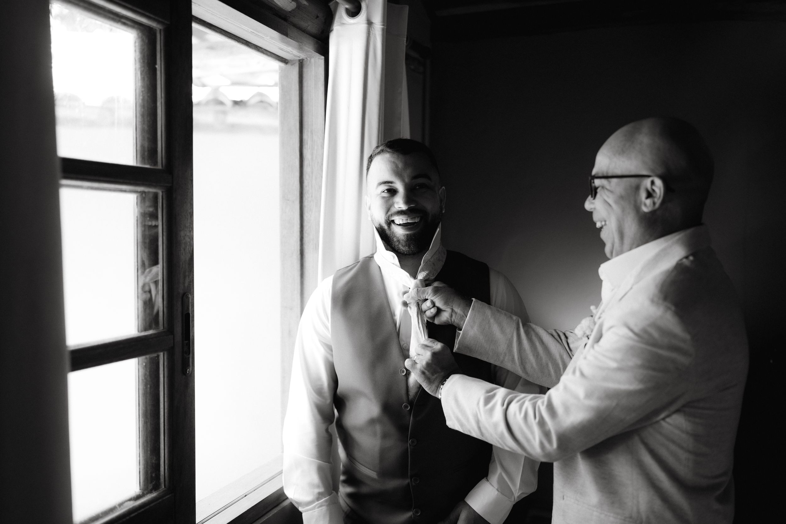 Two men in formal wear share a candid moment by a window in black and white.