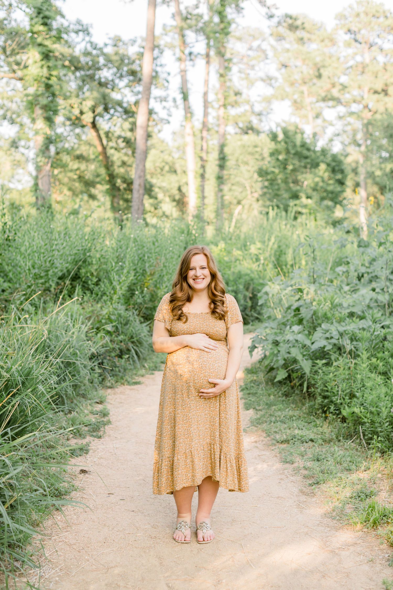 Pregnant woman in yellow dress stands alone on dirt path surrounded by greenery.