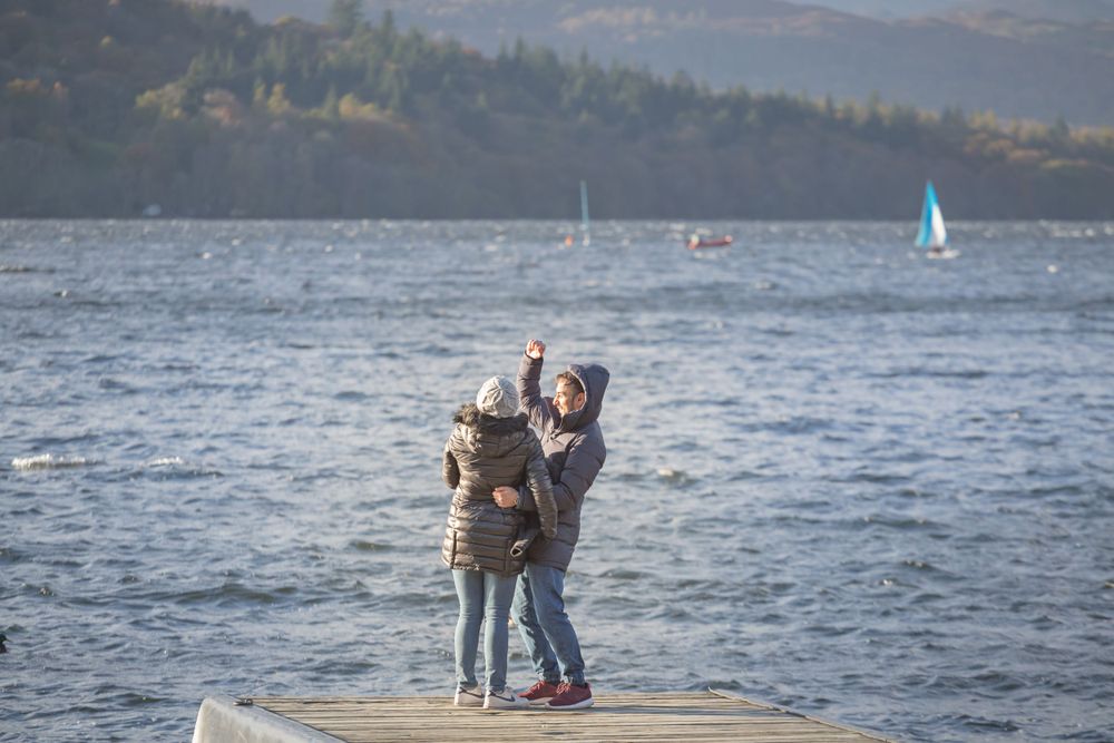 A pair stands close together on a dock with sailboats and mountains visible in the distance.