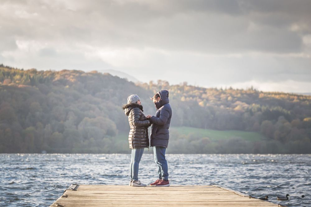 Two people share an intimate moment on a lakeside pier with autumn mountains in the background.