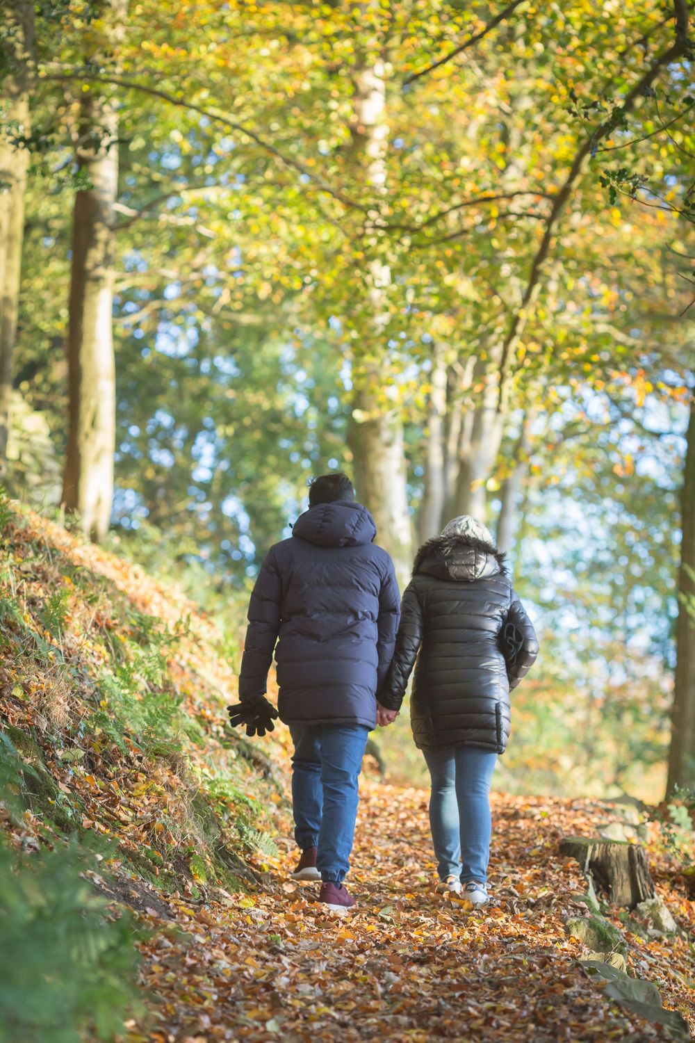 Two people in puffy winter coats walk along a leaf-covered forest trail in autumn.