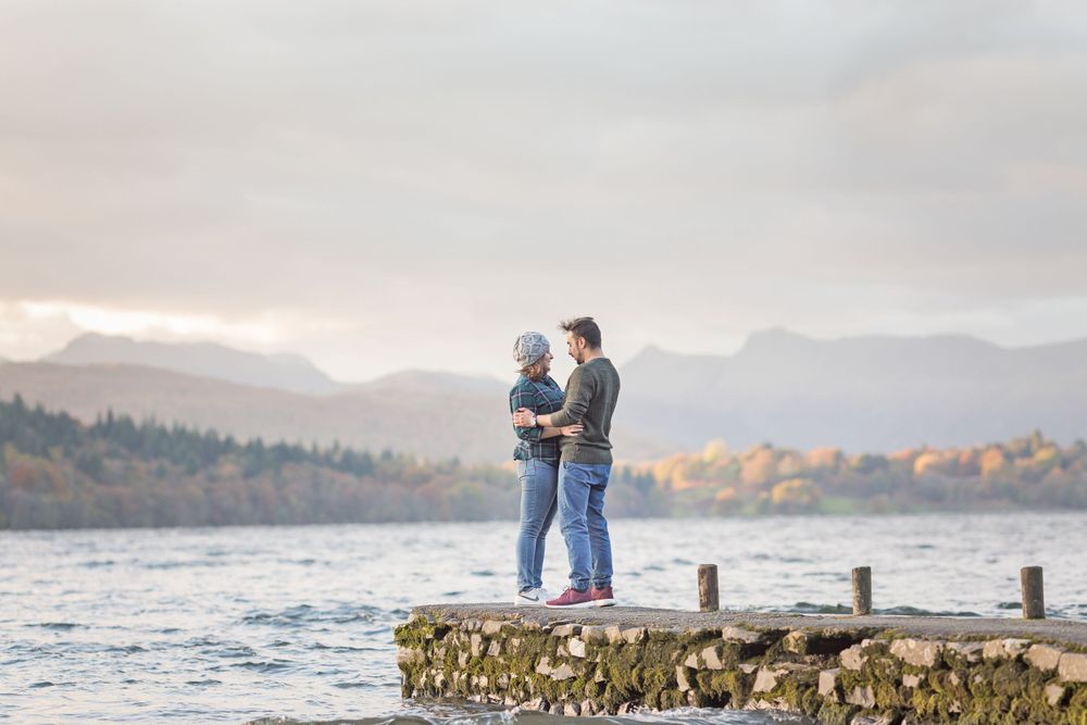 A couple stands together on a stone jetty overlooking a misty lake with mountains in the background.