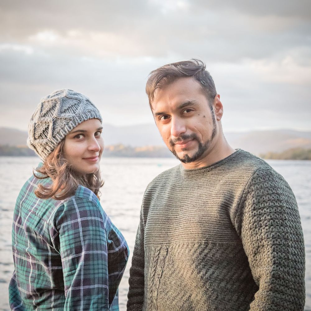 A couple in casual wear poses by a lake with mountains and cloudy skies in the background.