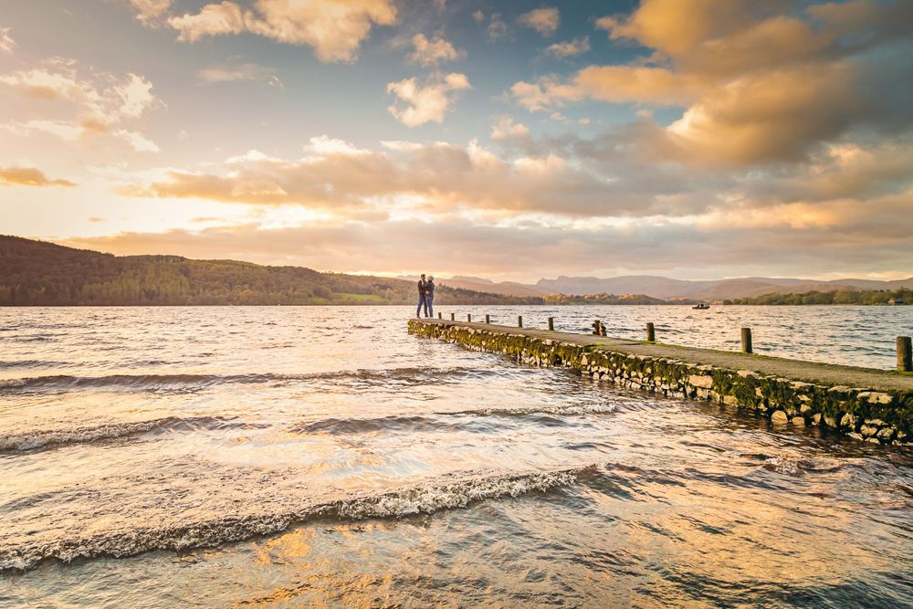 Stunning sunset over a lake with waves crashing against a stone pier and mountains in the distance.
