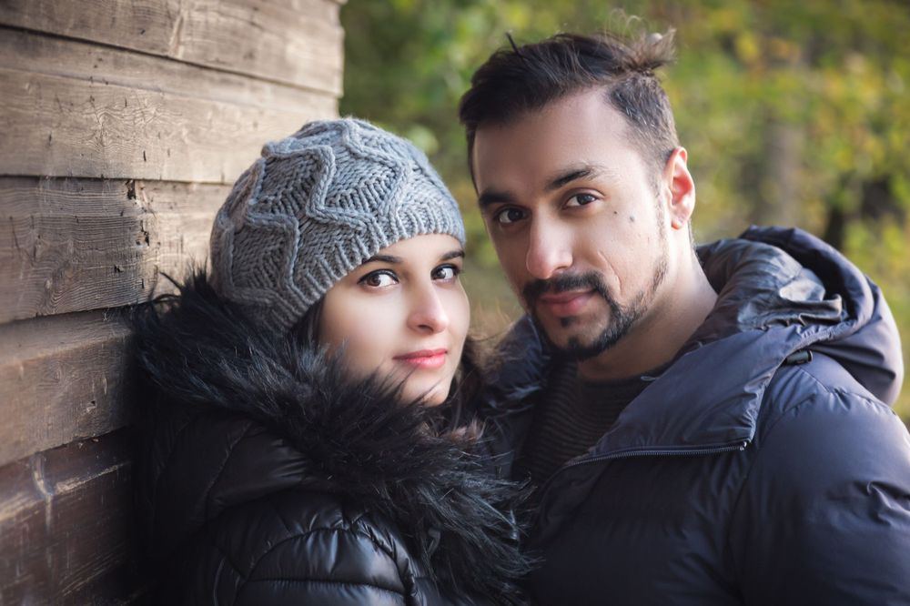 A couple in winter attire poses against a rustic wooden wall during an outdoor photoshoot.