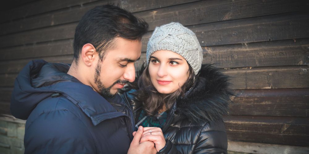 Romantic winter couple in puffy jackets and knit hat standing against wooden wall.