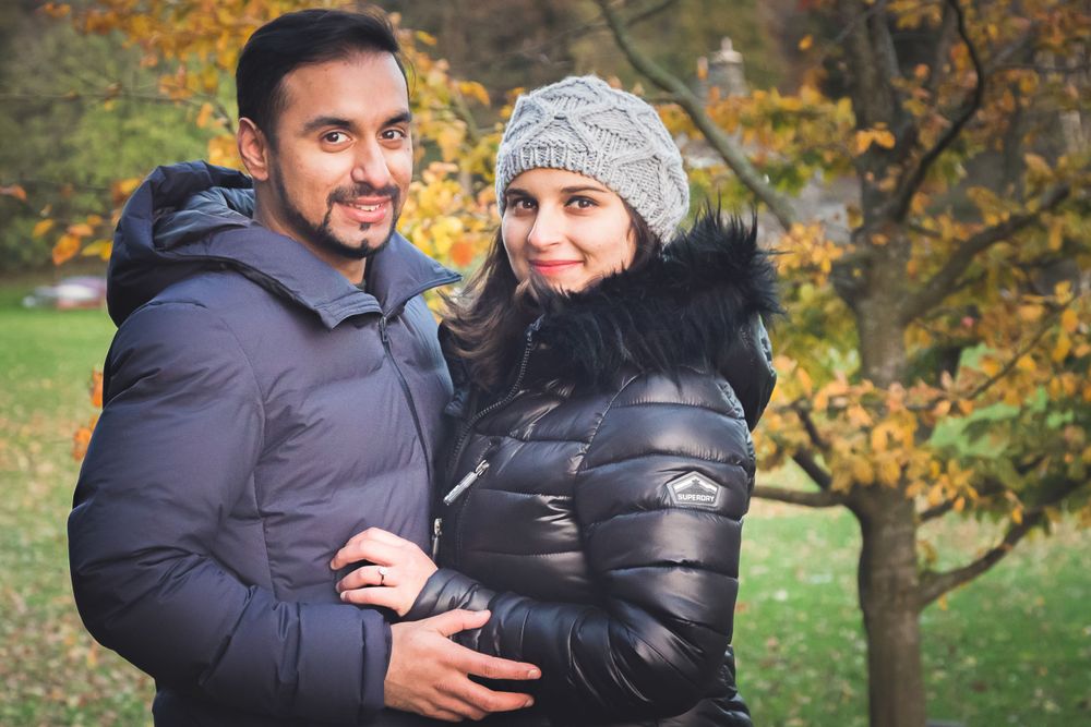 Happy couple in winter coats embracing outdoors with autumn foliage in background.