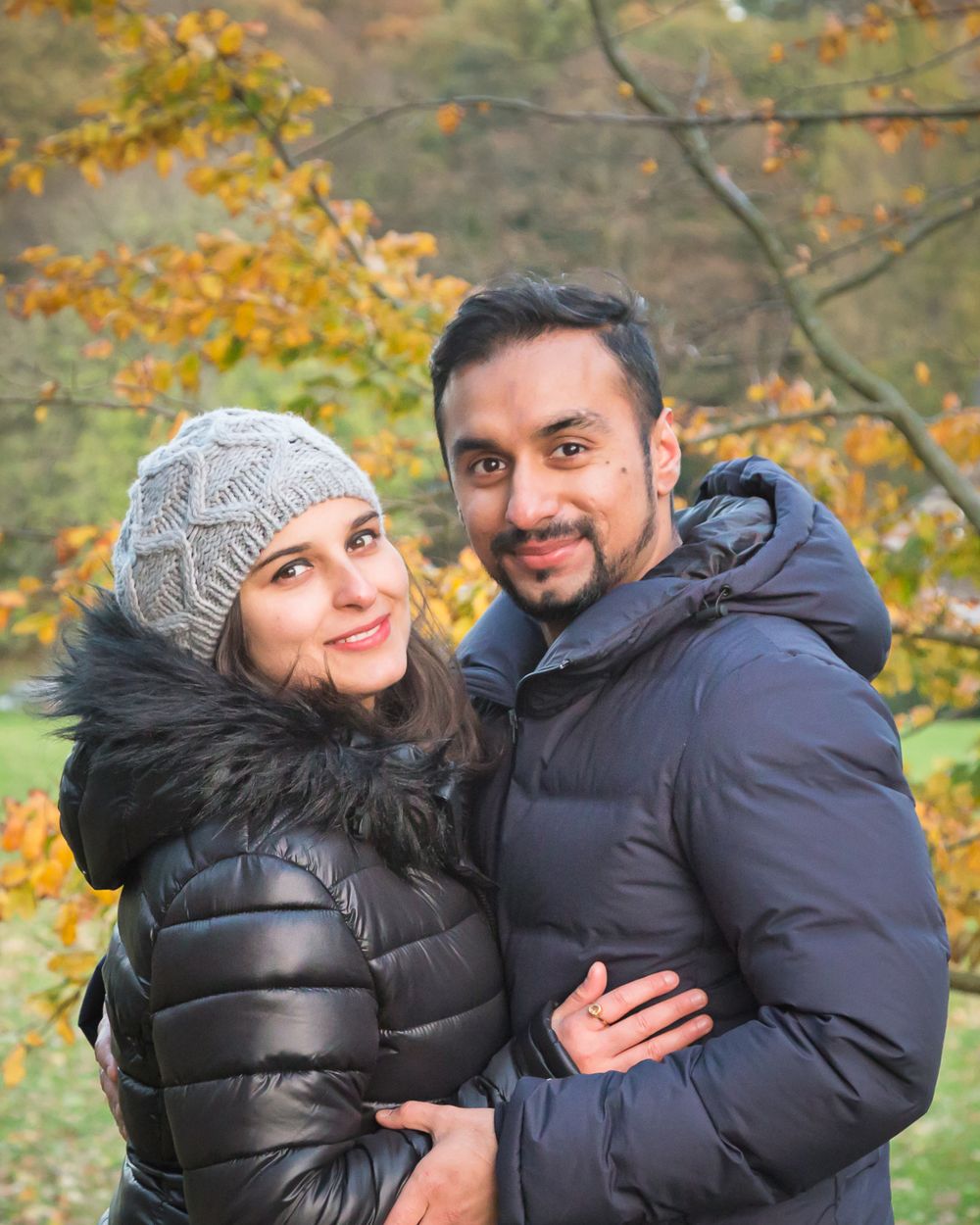 Smiling couple in winter coats pose together against colorful autumn trees.