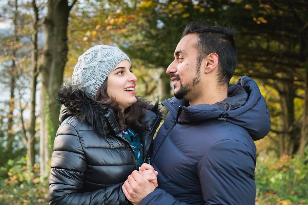 Candid moment of couple in winter wear smiling at each other in autumn forest.