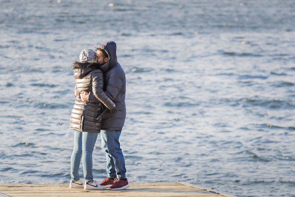 A couple embraces while standing on a wooden dock overlooking a serene lake on a cloudy day.