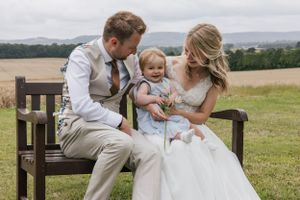 Abbie and Rob on their wedding day with their daughter sat on a bench in a field sharing a moment together and laughing
