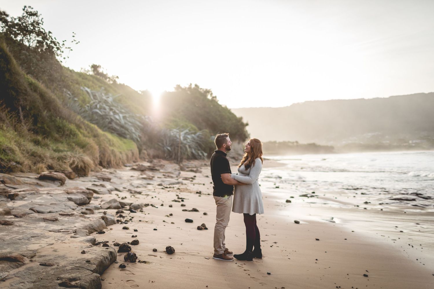 Couple embraces on rocky beach at sunset with mountains in background casting golden light across the shore.