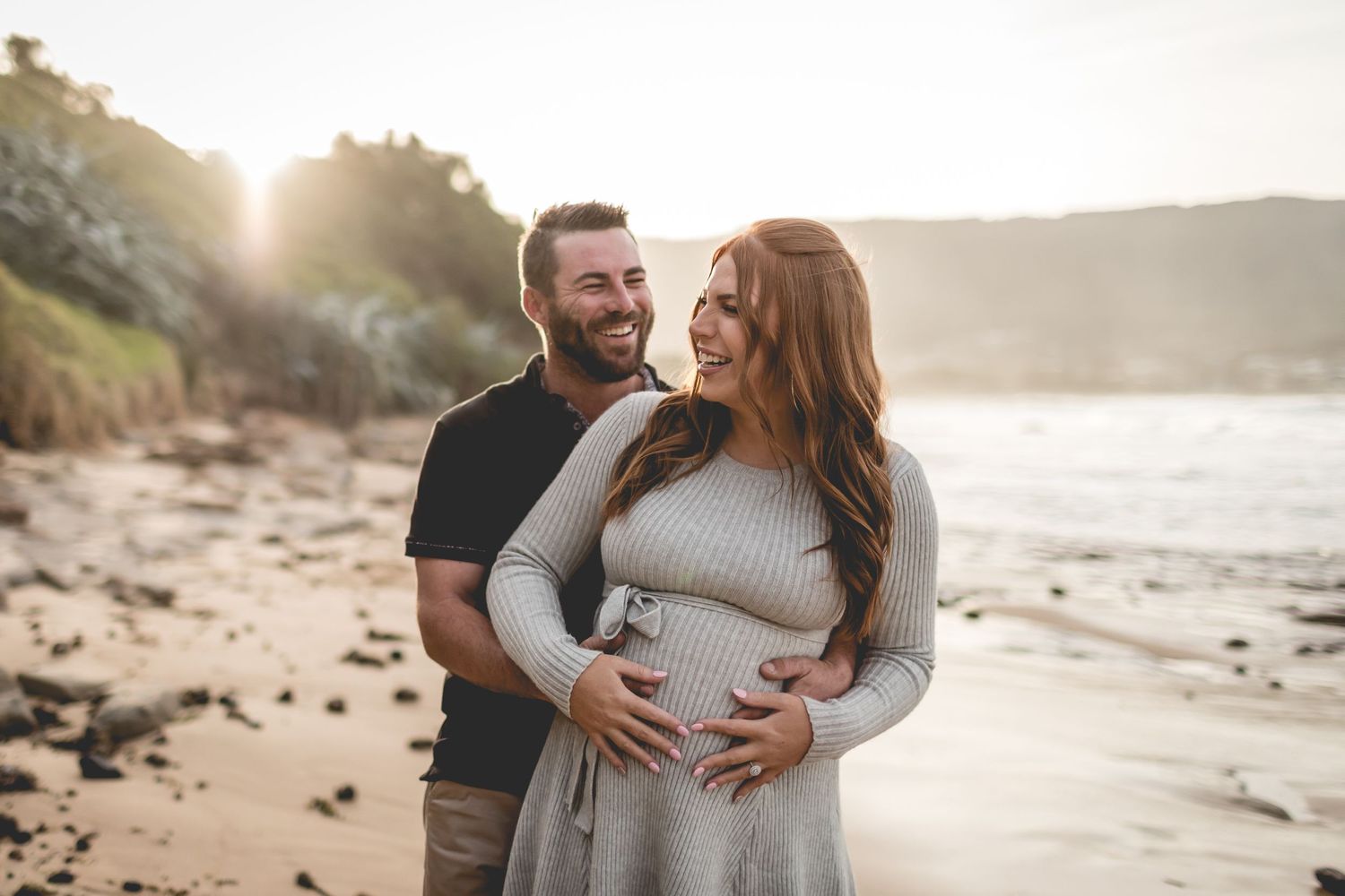 A couple embraces on a beach at sunset during their maternity photo session.