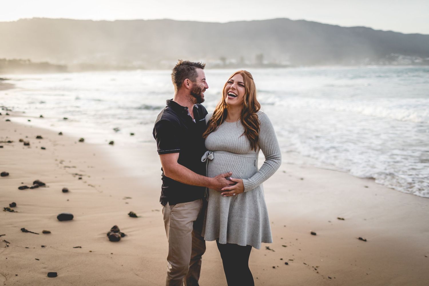 Couple shares tender moment on beach with waves and coastal mountains in background.