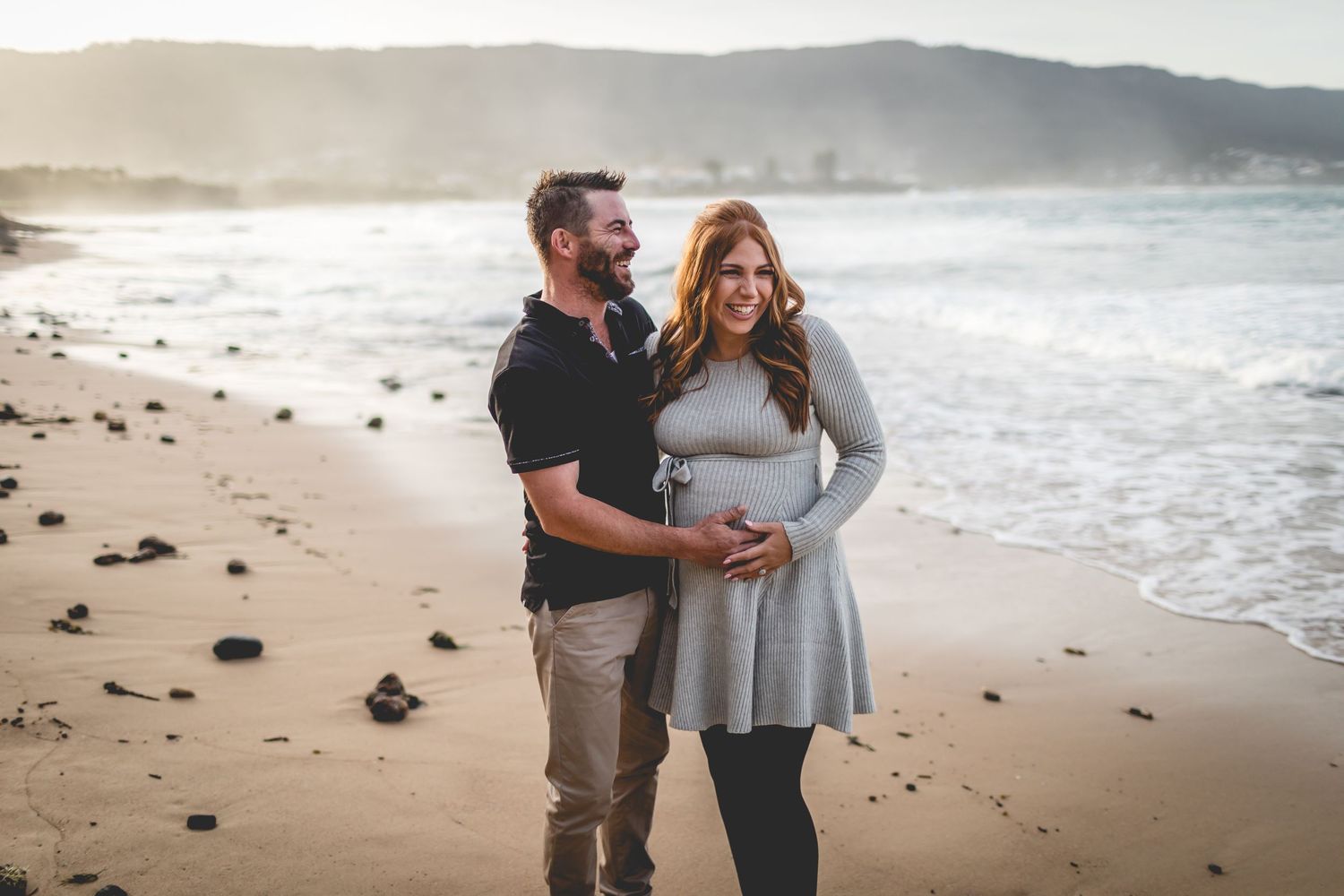 Couple stands together on rocky shoreline with misty coastal mountains and crashing waves behind them.