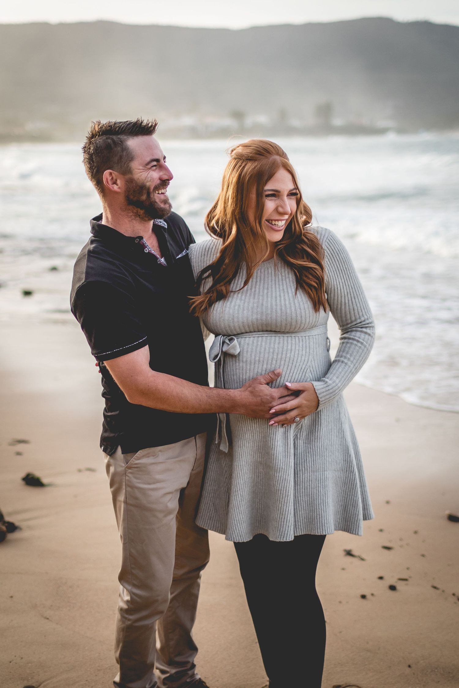 Couple shares joyful moment together on scenic beach with waves and mountains in background.