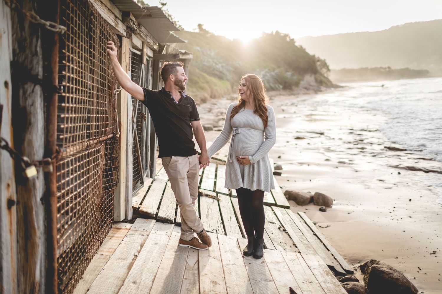 Couple stands together on wooden boardwalk along rocky coastline at sunset.