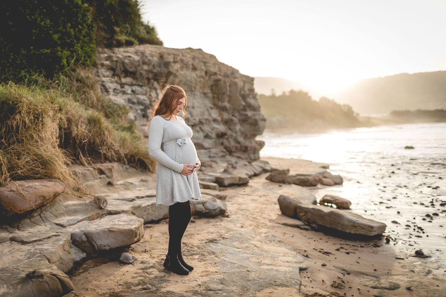 Person in gray dress stands on rocky coastal outcrop during golden hour with cliffs in background.