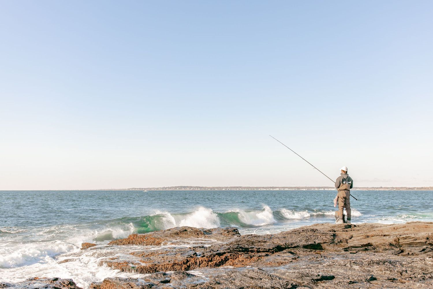 Lone fisherman casting a line from rocky shore with waves crashing beside him.