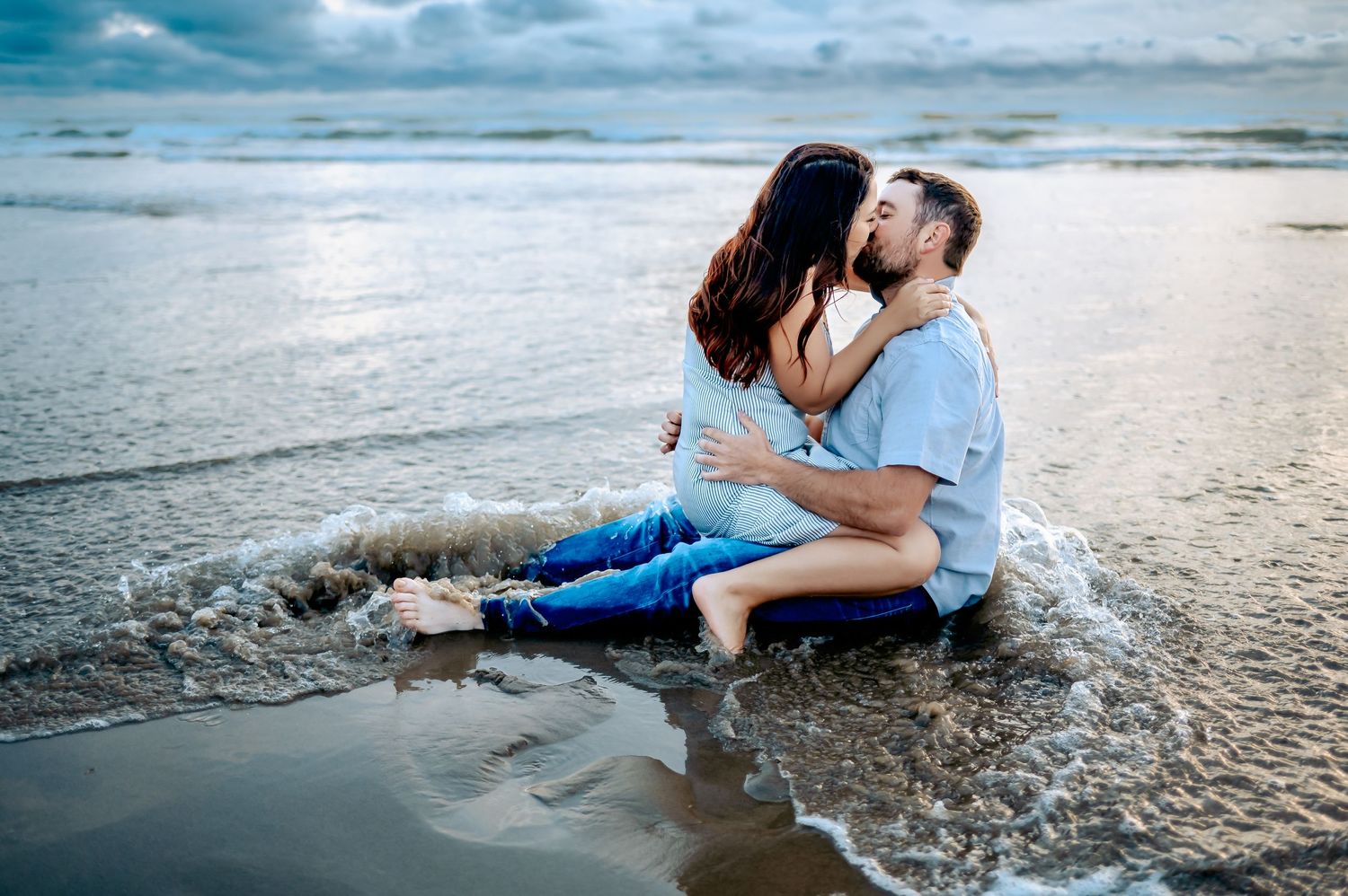 Couple sitting together on wet sand as ocean waves roll in during sunset.