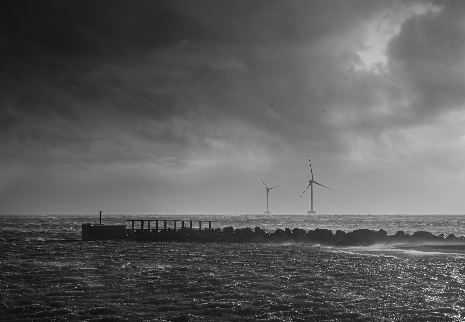 A dramatic black and white panoramic seascape with wind turbines and industrial structures along the horizon.