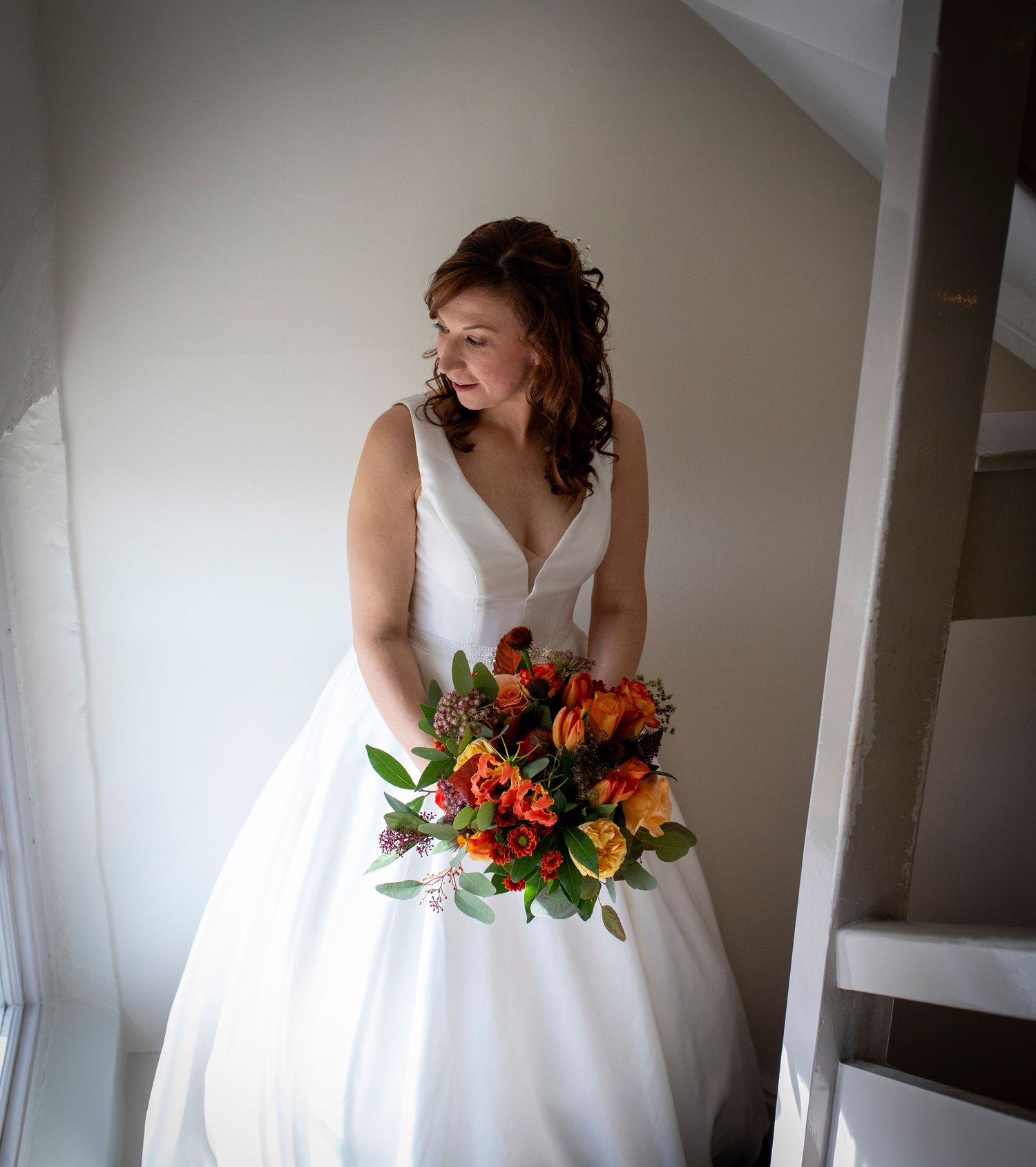 Bride in classic white gown holds vibrant orange and red bouquet near window light.