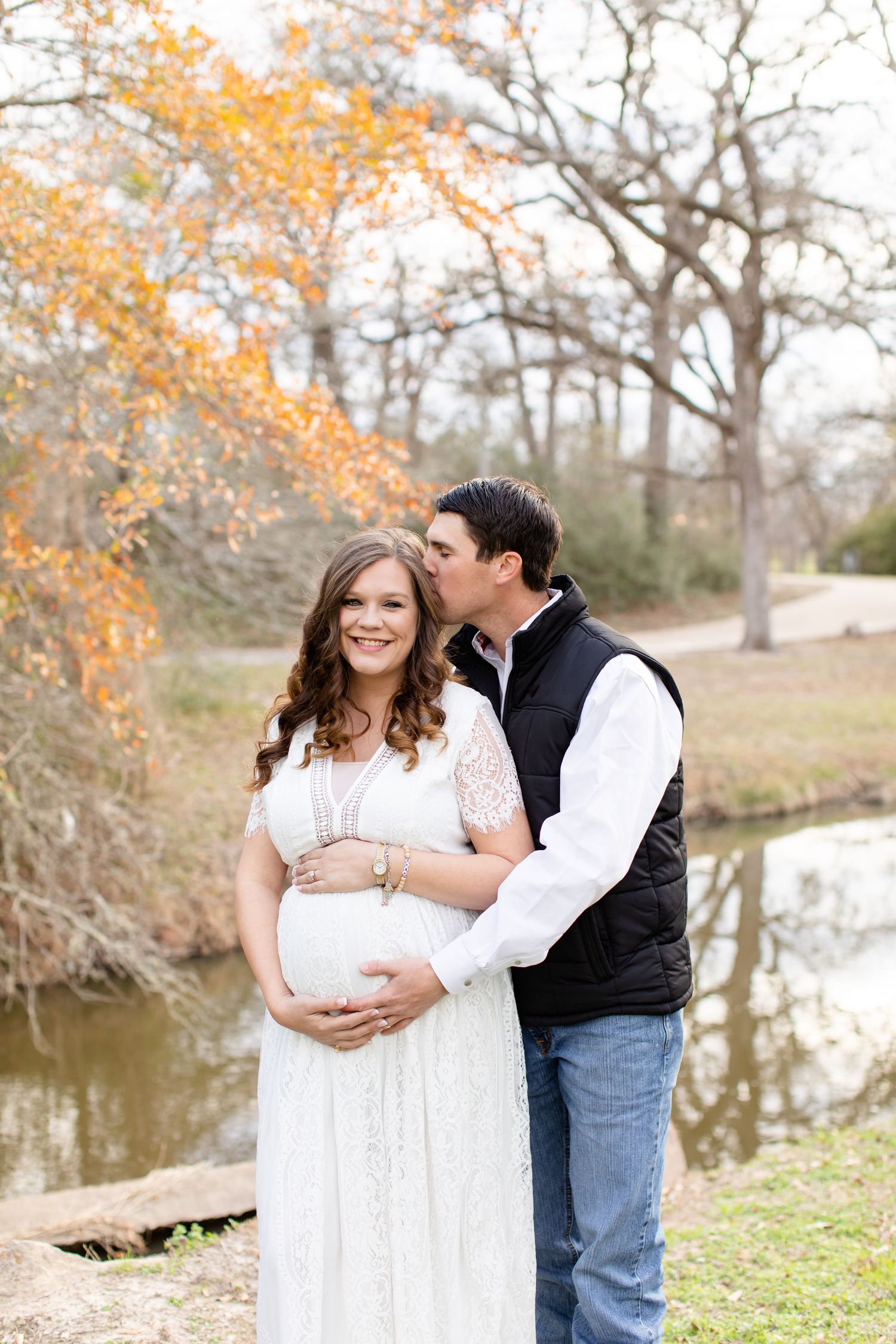 Maternity photo of couple by pond with fall foliage in background.
