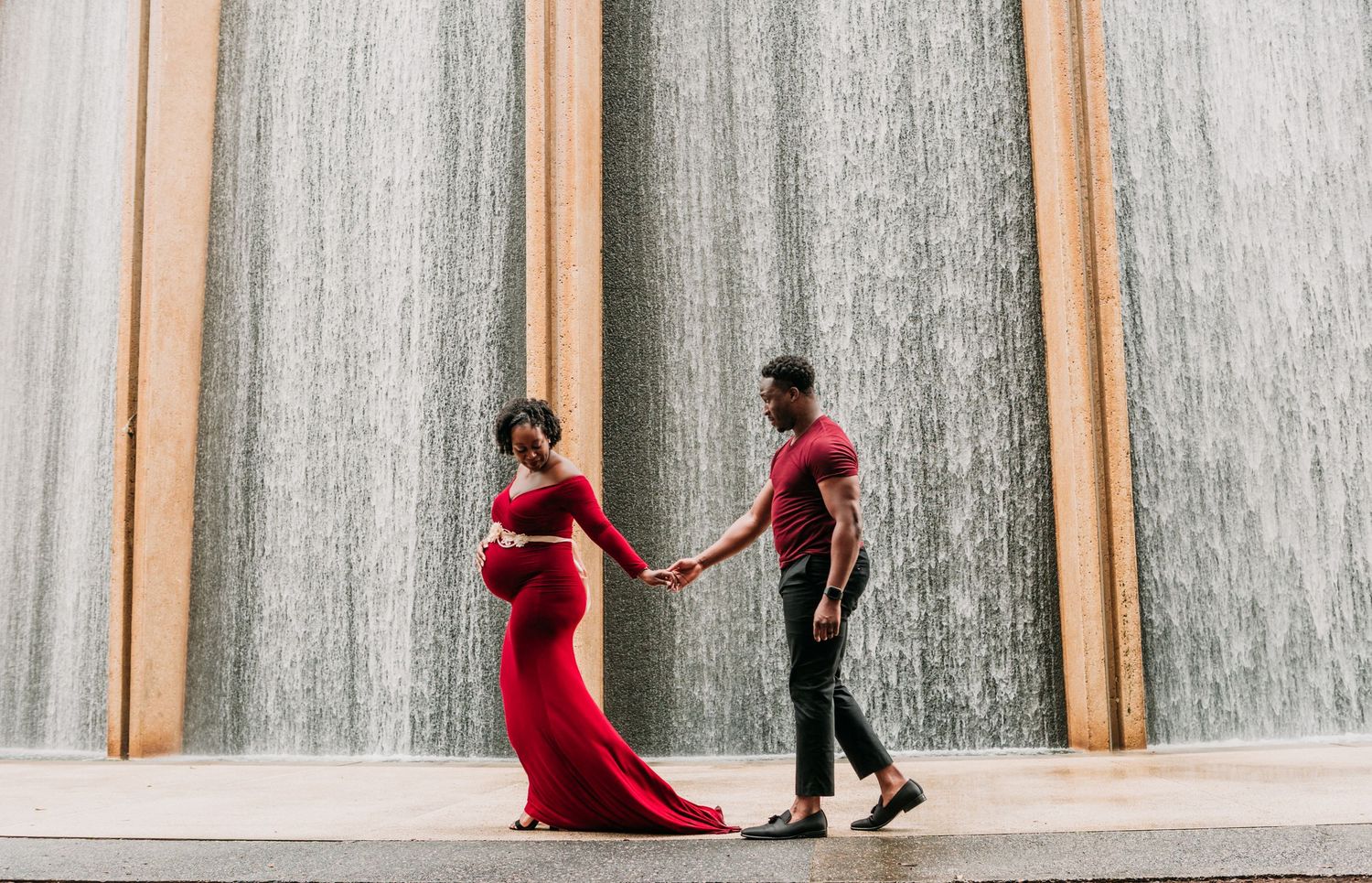 A couple holds hands against a dramatic cascading waterfall wall backdrop for a maternity session