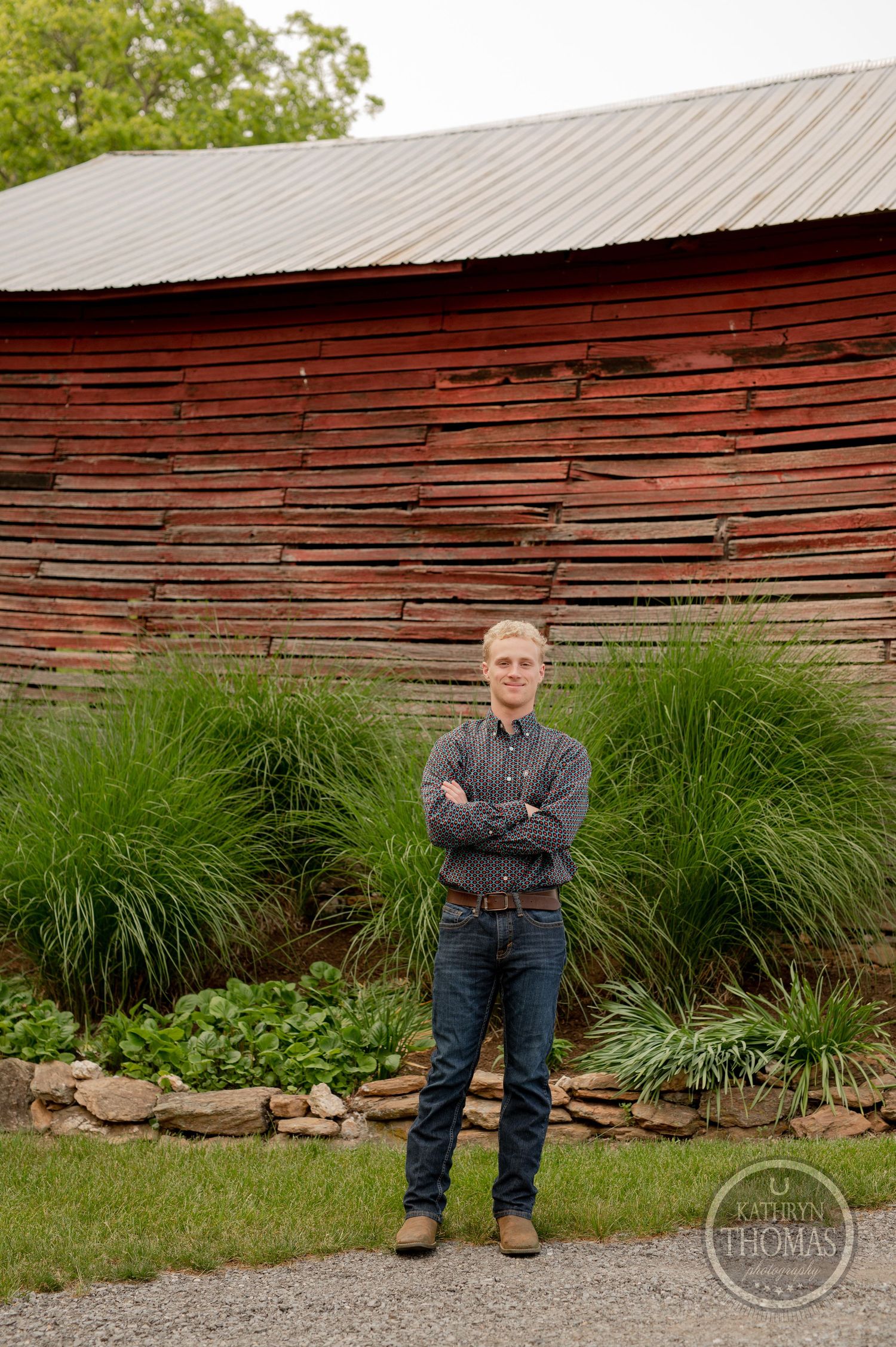 Person in denim outfit stands confidently in front of rustic red barn with landscaped garden.