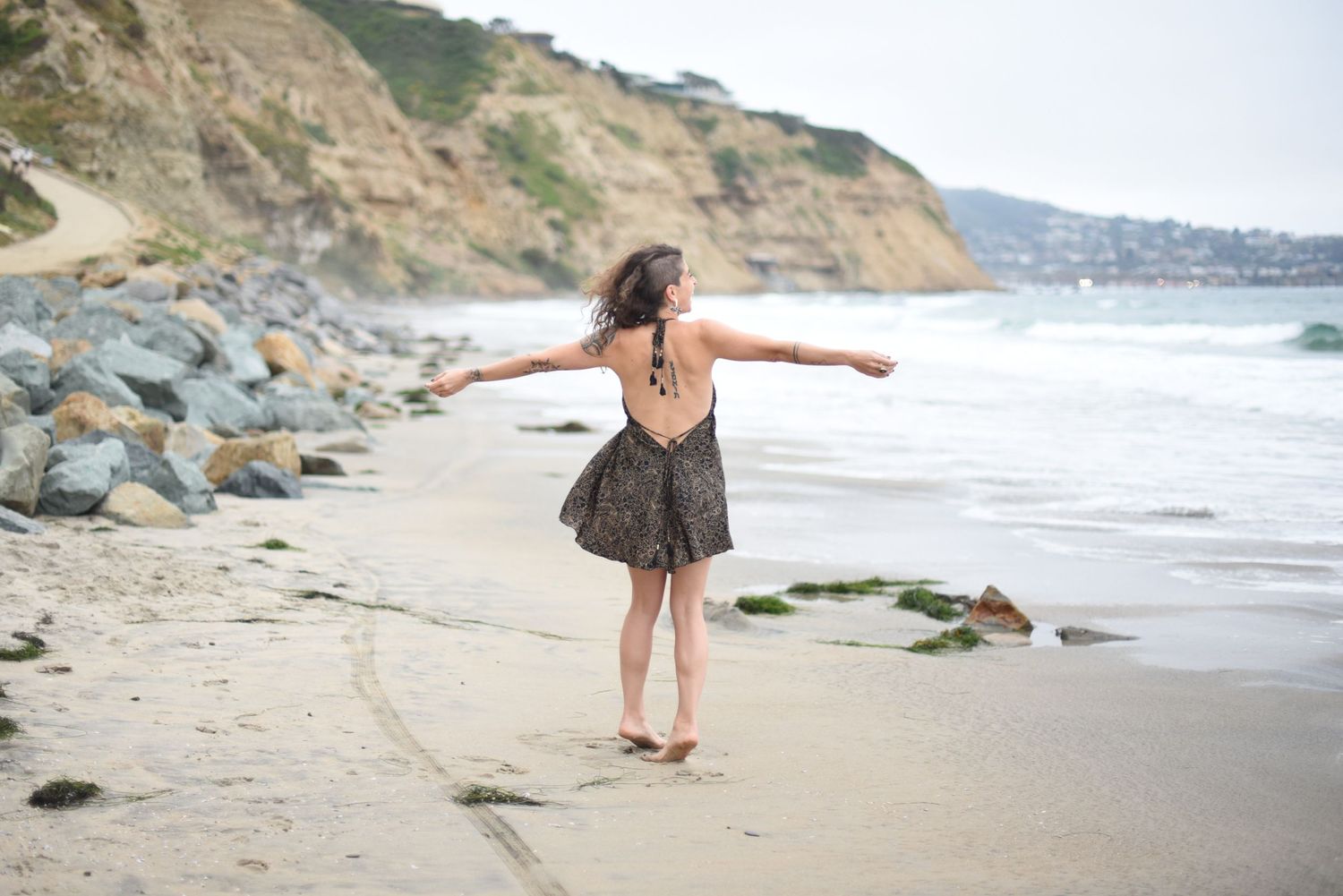 A figure in a dress stands with arms outstretched on a rocky beach.