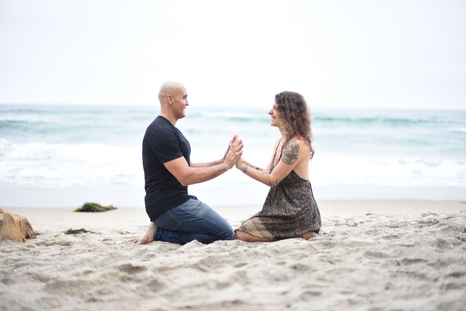 Two people sit facing each other holding hands on a sandy beach.