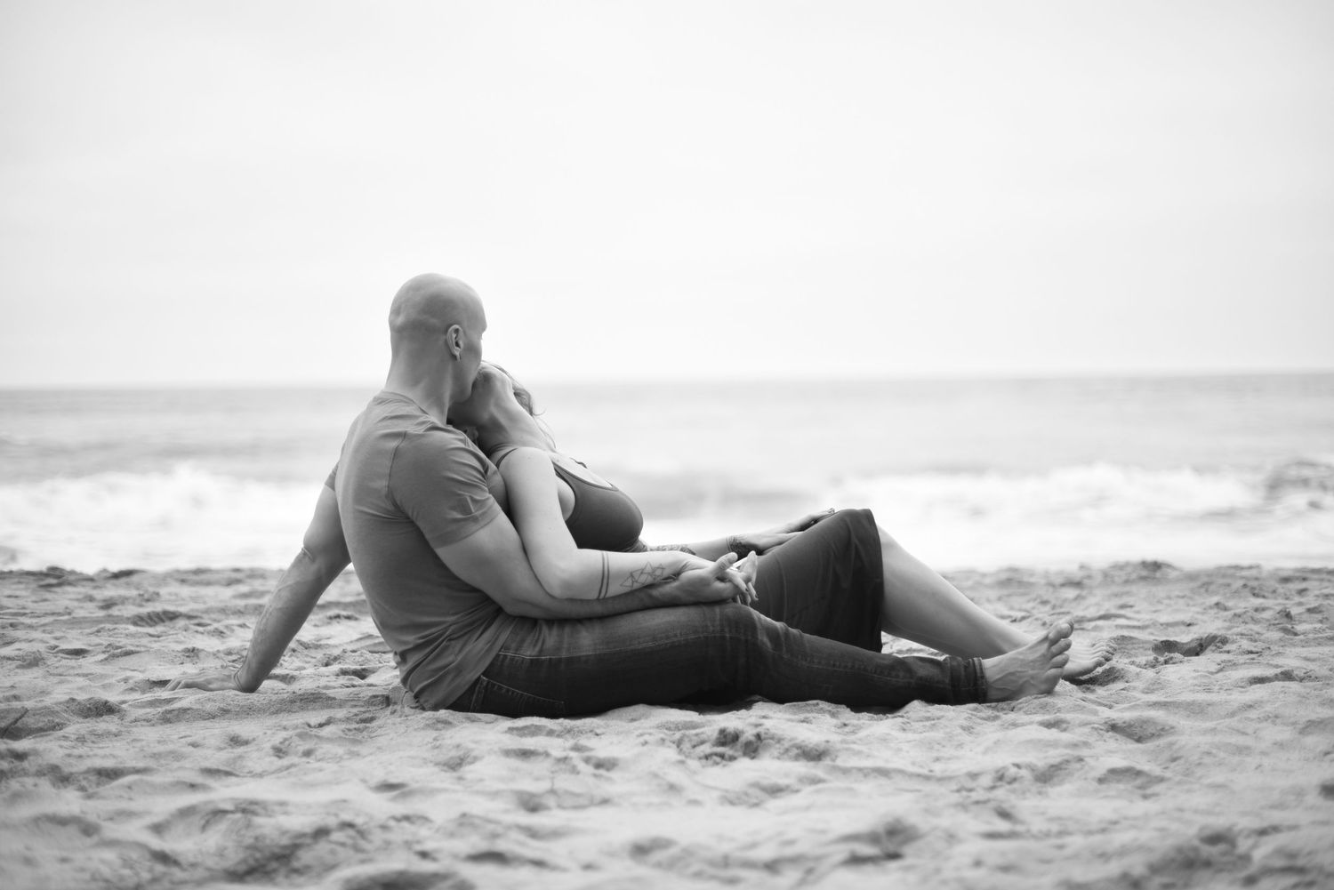 A couple relaxes intimately together on a sandy beach with waves in the background.