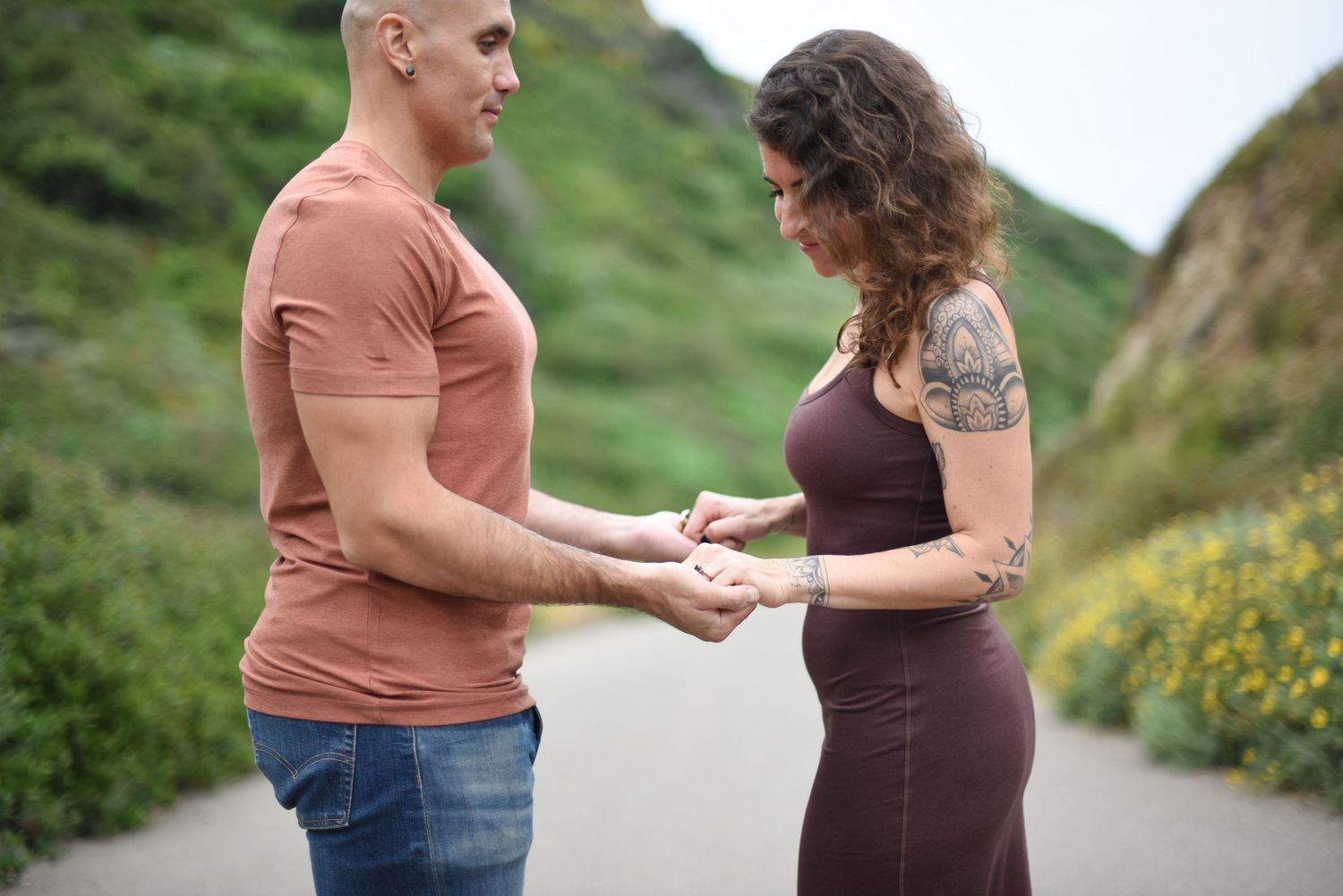 Couple in casual wear holding hands on coastal path surrounded by yellow wildflowers.