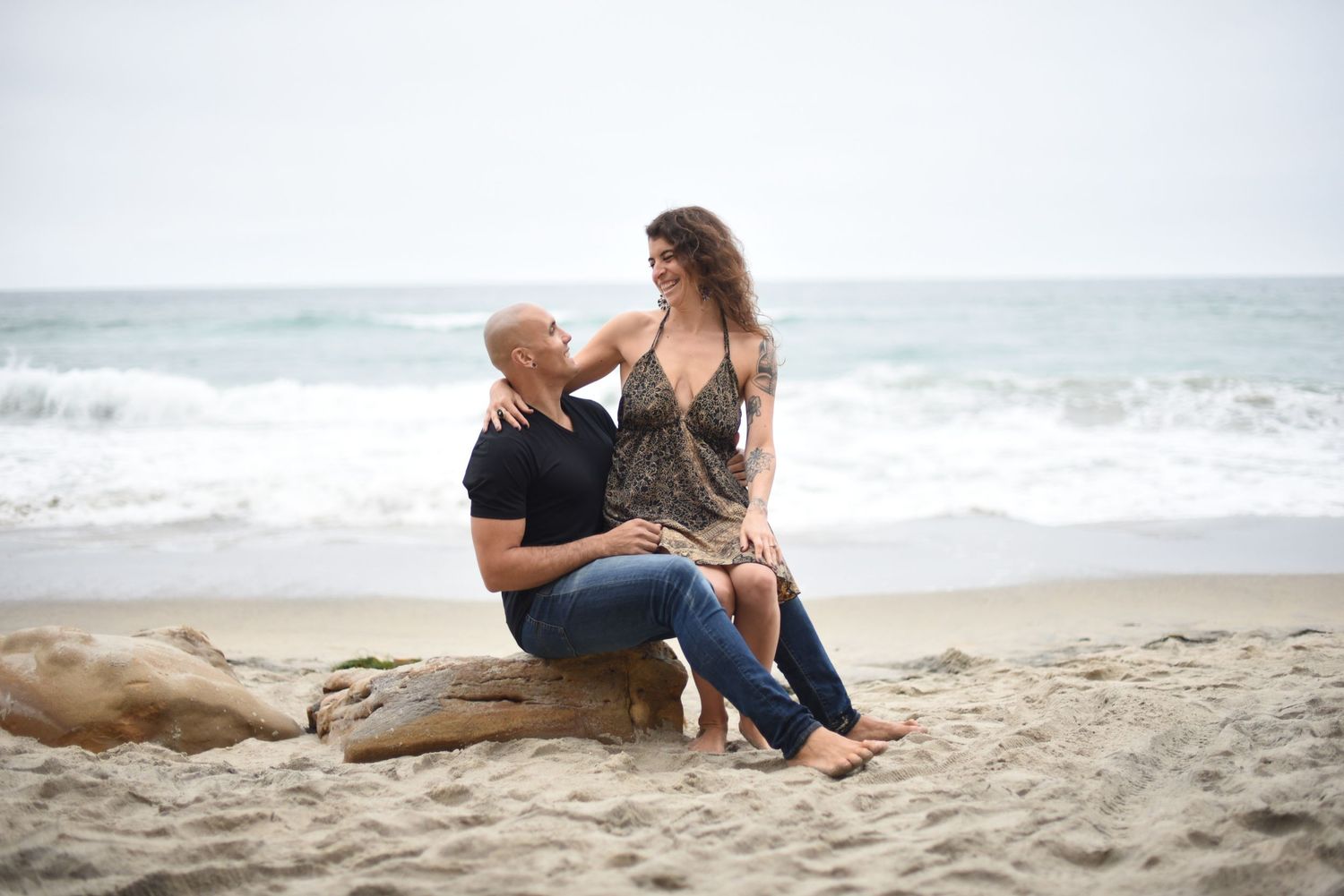 A couple shares a tender moment while sitting on driftwood at the beach.