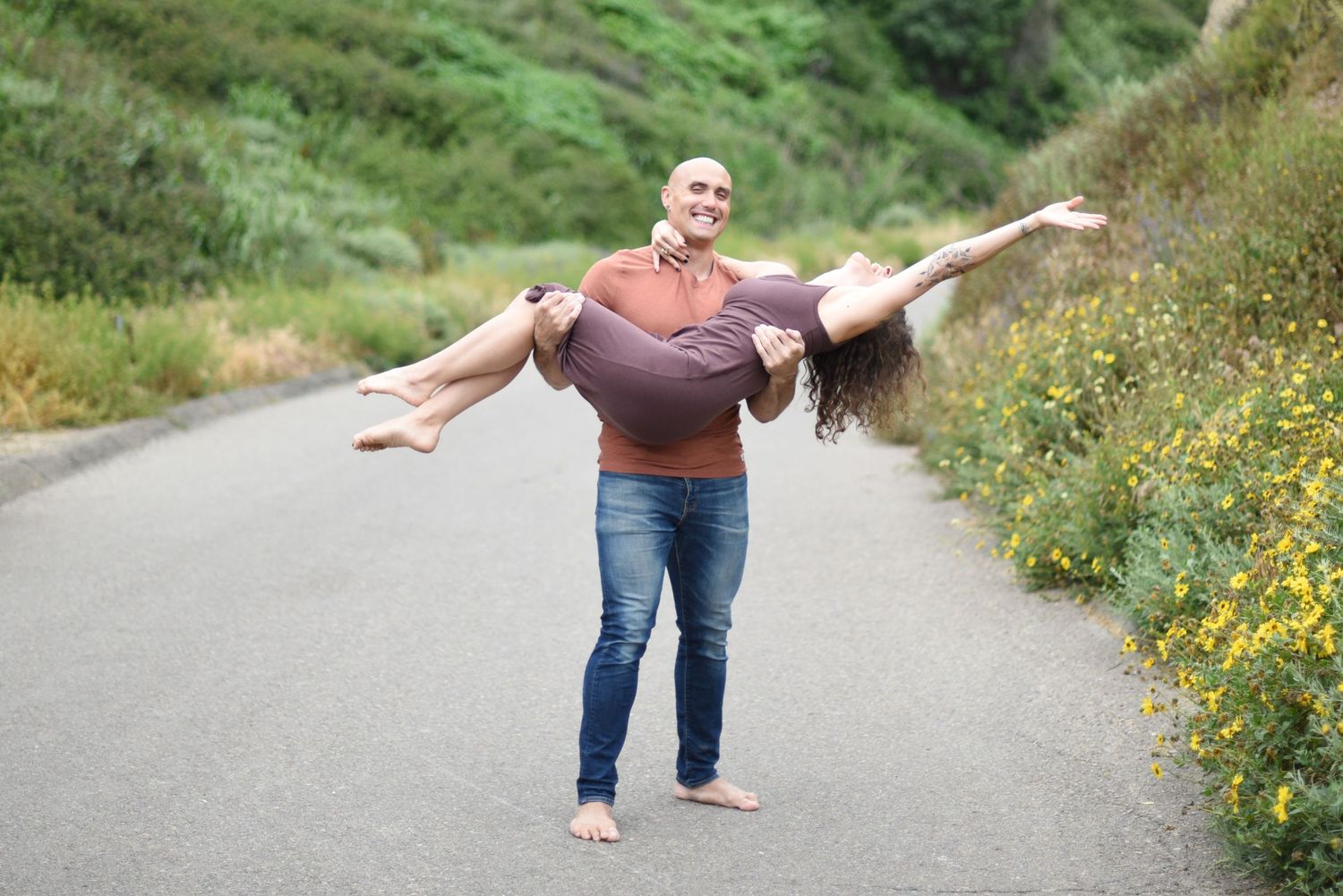 Playful couple on mountain path with dramatic lift pose among wildflowers.